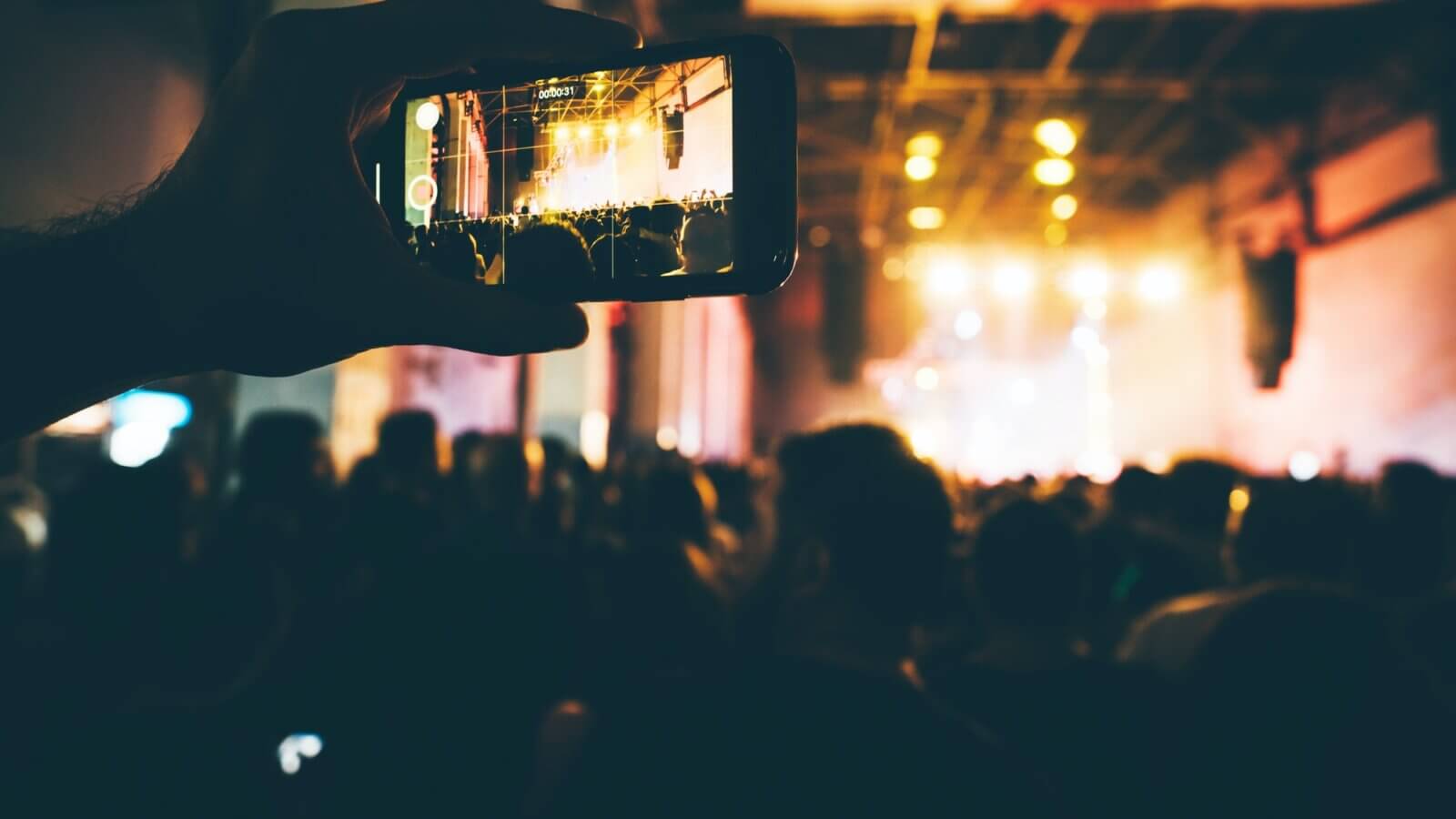 Close up hand with phone at the concert hall. Crowd at the concert and blurred stage lights.