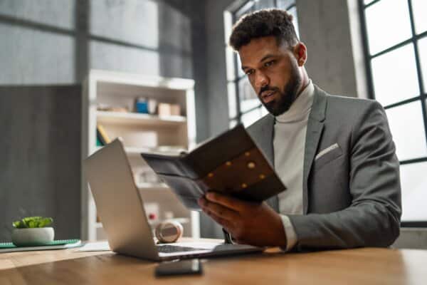Young african american businessman with diary working on laptop indoors in office