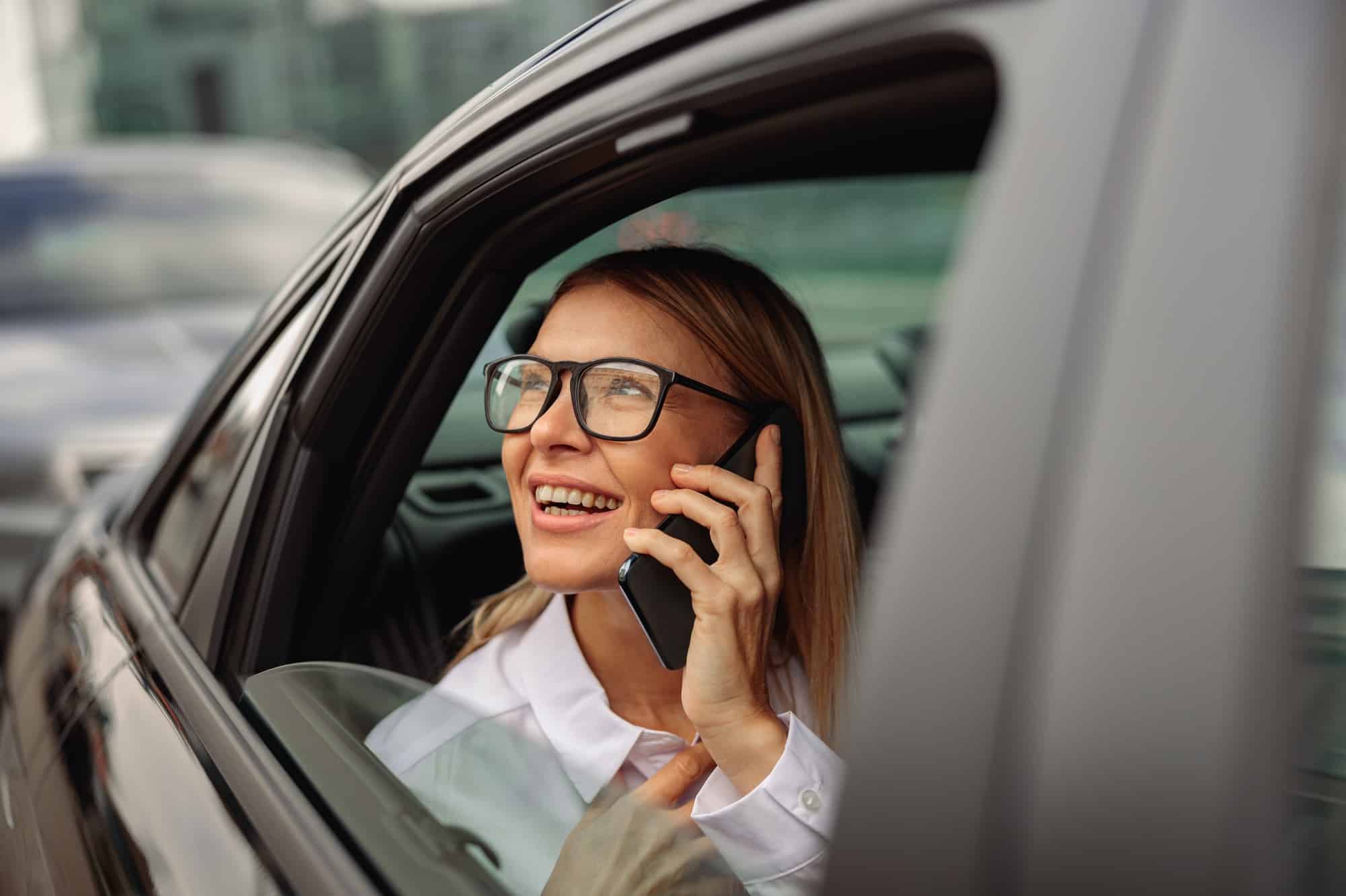 Happy woman in eyeglasses traveling on back seat of car and talking smartphone while looking